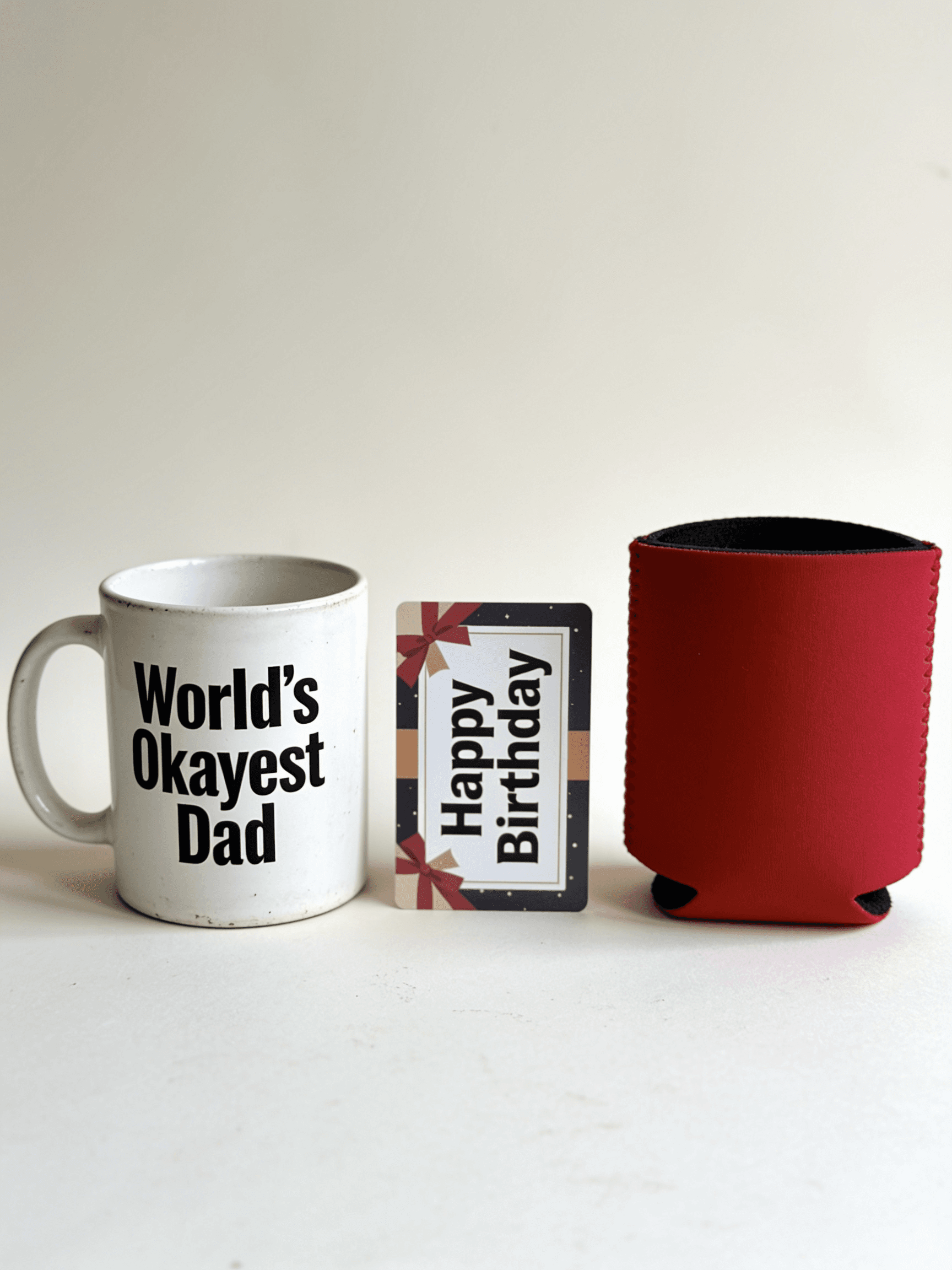 The lineup of polite, forgettable dad gifts: a 'World's Okayest Dad' mug, a Happy Birthday gift card, and a red beer koozie